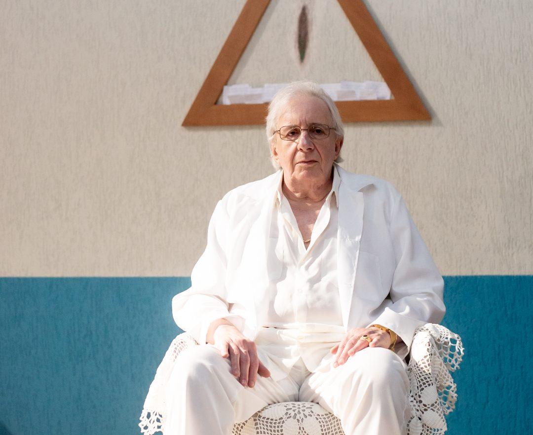 o ator marco nanini sentado como joão de deus, ele se veste todo de branco e olha pra a câmera. Imagem da série João Sem Deus.