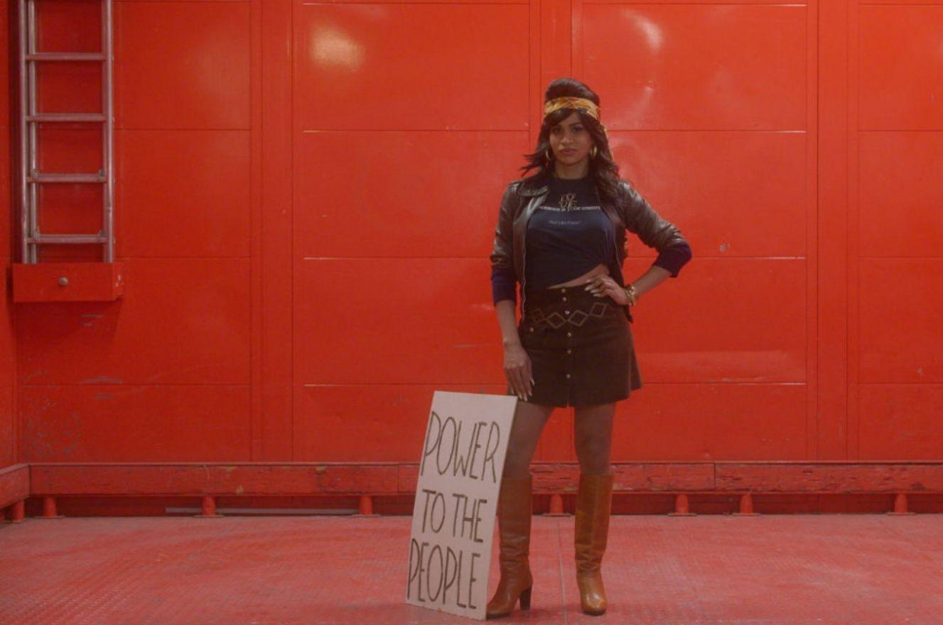 Mulher trans com a mão na cintura, de pé, olha para a câmera. A parede ao fundo é vermelha. É uma cena do filme Orlando.