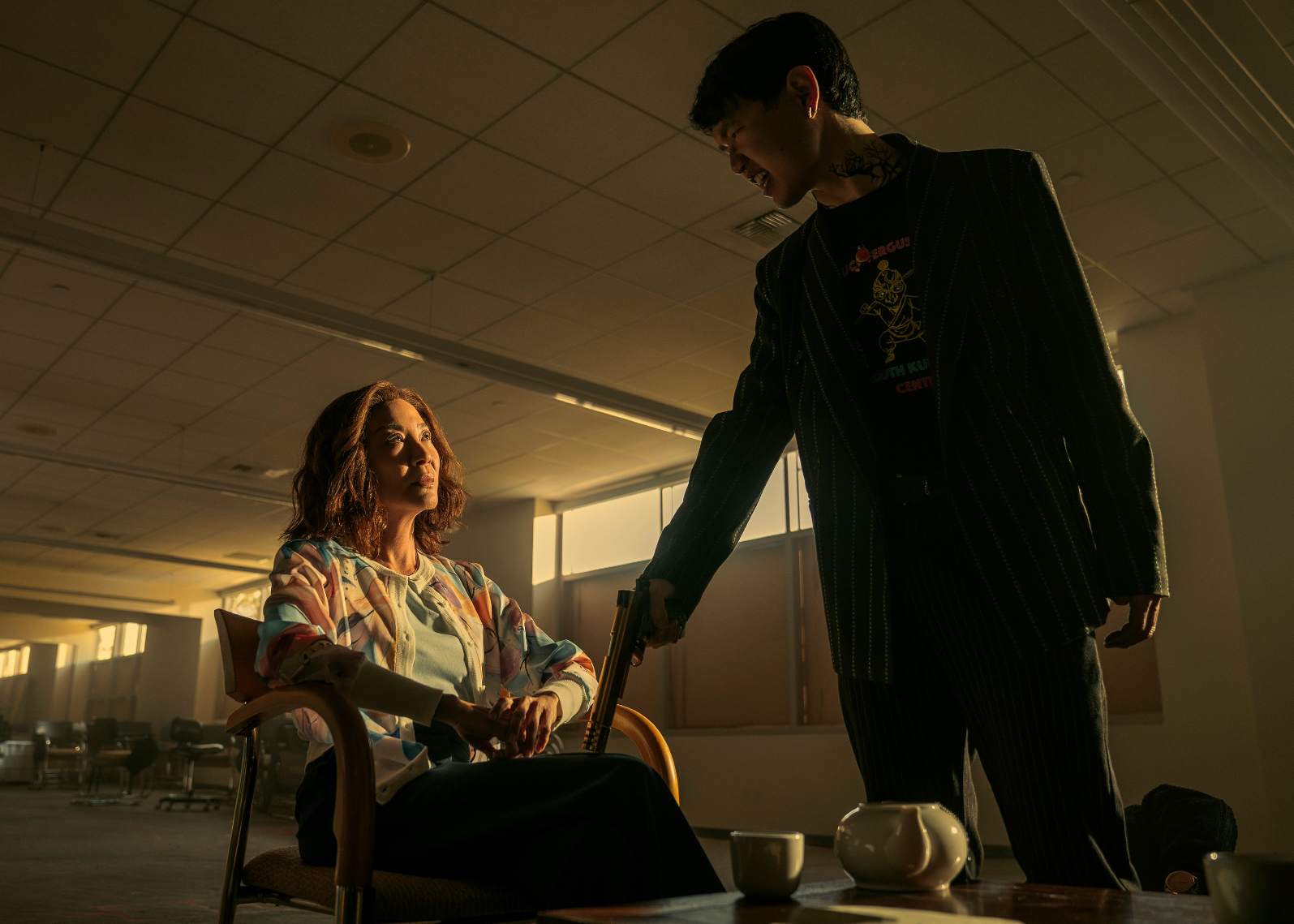 Cena de Irmãos Sun . Michelle Yeoh sentada, enquanto um bandido asiático aponta arma pra sua perna.